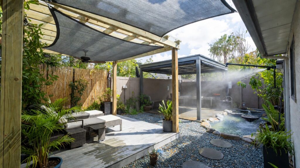 outdoor living 3 Backyard patio designed for humid weather comfort: wooden pergola with dark shade sail for ventilation, ceiling fan, misting system creating cooling fog, small pond and fountain, surrounded by tropical plants and moisture-resistant composite decking and gravel.